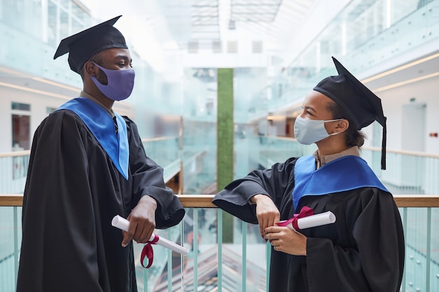 Retrato lateral de dois jovens afro-americanos usando batas de formatura e máscaras enquanto conversam em ambientes fechados