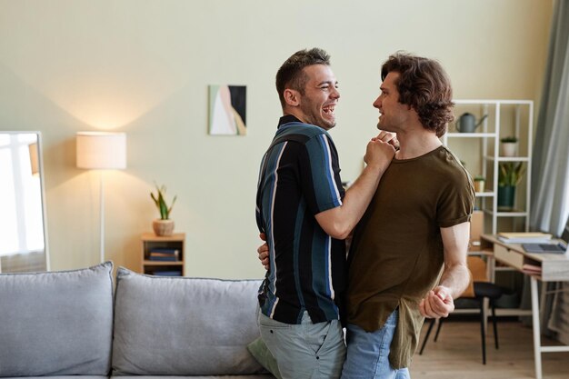 Retrato lateral de um casal gay despreocupado dançando na nova casa e rindo alegremente copiando o espaço