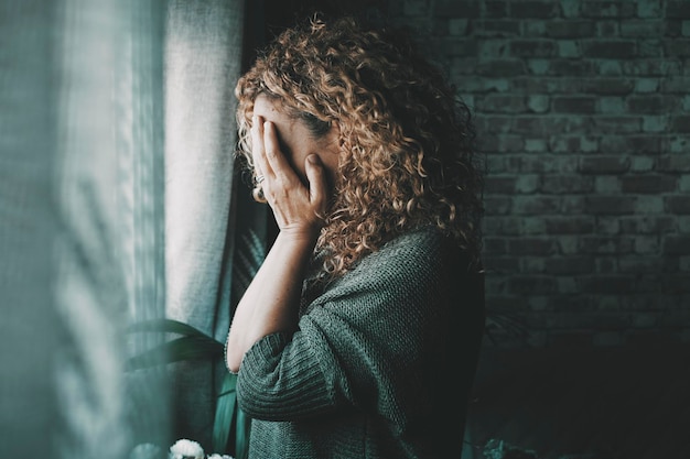 Retrato lateral de mujer triste y preocupada en casa en la oscuridad mirando por la ventana Mujer sola preocupada y solitaria interior Concepto de fin de relación y emoción fallida Señora tocando la cara