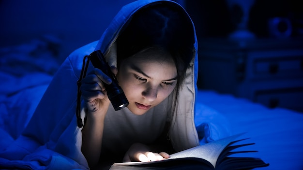 Retrato de libro de lectura adolescente en la cama con linterna.