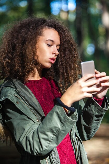 Retrato de la linda adolescente de cabello rizado con teléfono inteligente en el bosque de otoño