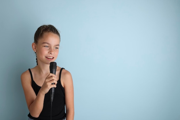 Retrato de una linda adolescente cantando con micrófono Adolescente con camiseta negra sobre un fondo azul aislado Karaoke para niños Entretenimiento en el hogar para niños Cantante cantando con micrófono