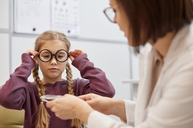 Retrato de linda adolescente poniéndose gafas grandes durante la prueba de la vista en la clínica de oftalmología moderna