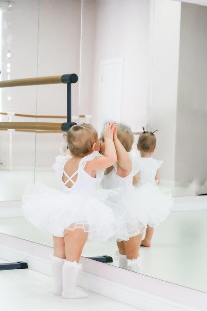 Retrato de una linda bailarina bebé sentada cerca de la barra de ballet poniéndose zapatos