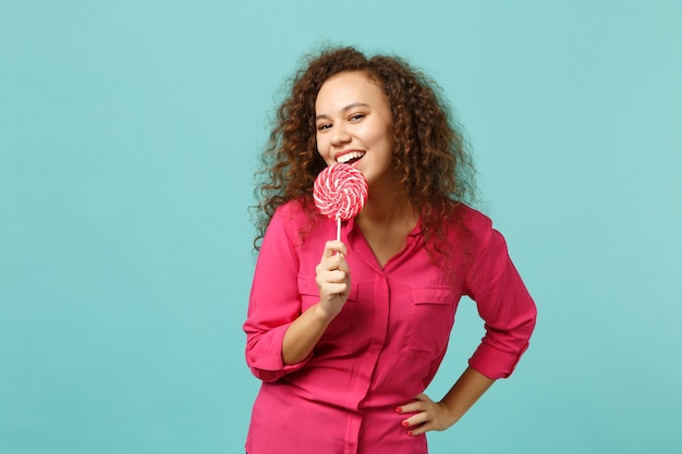 Retrato de linda chica africana en ropa casual sostenga en la mano comiendo piruleta redonda rosa aislada sobre fondo azul turquesa en estudio. Personas sinceras emociones, concepto de estilo de vida. Simulacros de espacio de copia.