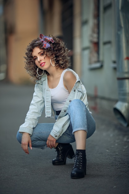 Retrato de una linda chica al estilo de los 90. Una niña con una camiseta blanca y jeans está en cuclillas. Modelo con rizos en la cabeza y lápiz labial rosa en el fondo de un paisaje urbano.