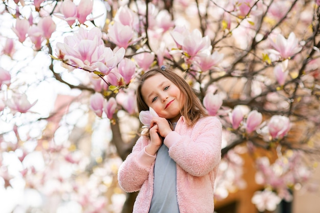 Retrato de una linda chica alegre con un vestido gris con chaqueta rosa en primavera en un parque bajo un árbol de magnolia rosa en flor.