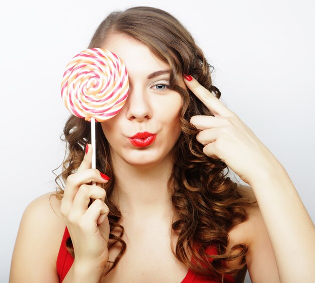 Retrato de una linda chica sonriente que cubre su ojo con piruleta sobre fondo blanco.