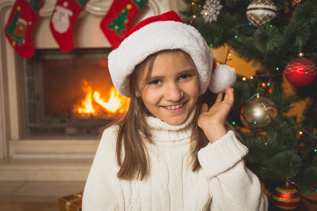 Retrato de linda chica en suéter blanco y gorro de Papá Noel posando en la chimenea en la sala de estar
