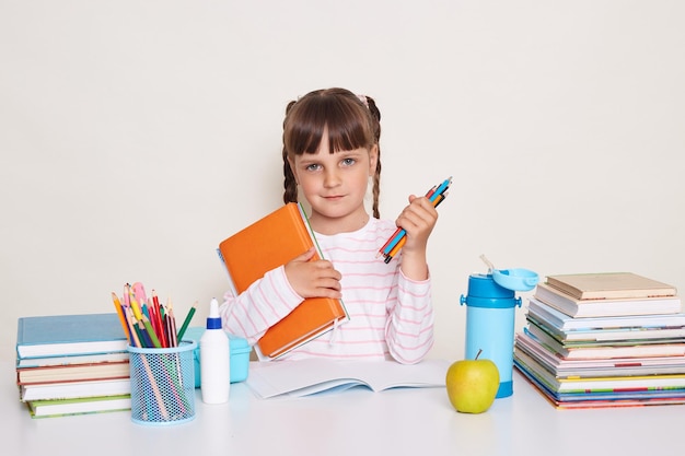 Retrato de una linda colegiala encantadora con cabello oscuro y trenzas sentada en la mesa sosteniendo un libro y lápices mirando a la cámara esperando a su maestra y comenzando la lección