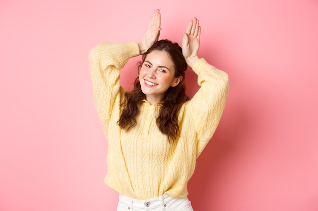 Retrato de linda dama mostrando gesto de orejas de conejo de Pascua y sonriendo posando como conejo tonto de pie contra la pared rosa