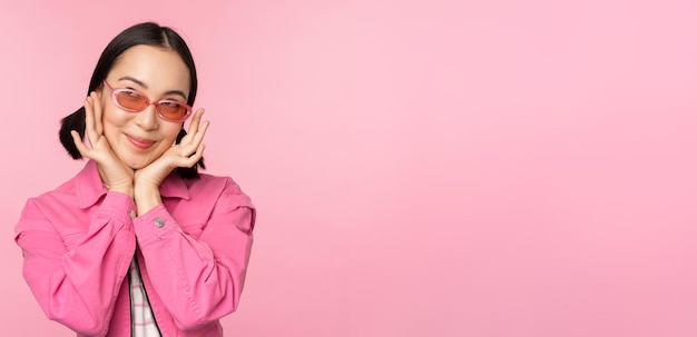 Retrato de una linda y elegante chica asiática sonriendo y tocando la cara mirando hacia arriba con una mirada pensativa de ensueño de pie sobre un fondo rosa