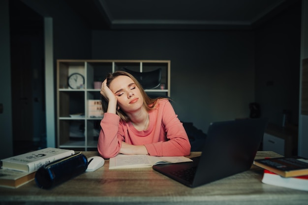 Retrato de una linda estudiante cansada con suéter rosa durmiendo en el escritorio en casa cerca de una laptop