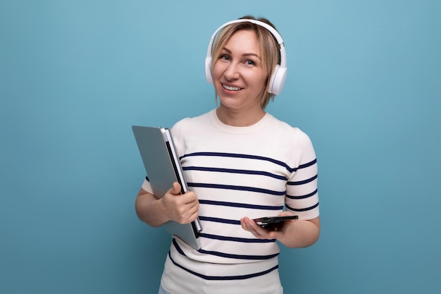 Retrato de linda estudiante rubia sonriente con grandes auriculares inalámbricos y portátil en azul