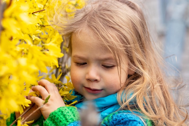 Retrato de una linda y feliz niña rubia con chaqueta azul cerca de un arbusto que florece con flores amarillas