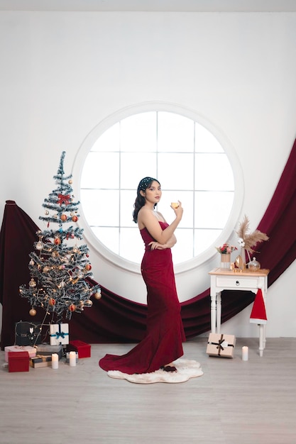 Retrato de una linda joven con un vestido rojo y bebiendo vino sonriendo a la cámara de pie en un salón de Navidad decorado en el interior