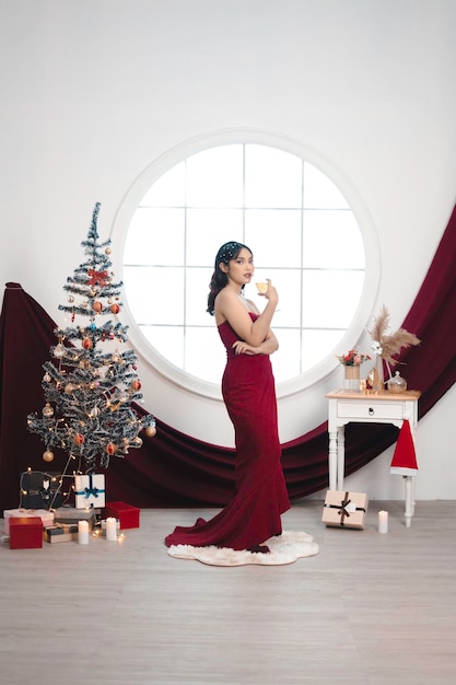 Retrato de una linda joven con un vestido rojo y bebiendo vino sonriendo a la cámara de pie en un salón de Navidad decorado en el interior