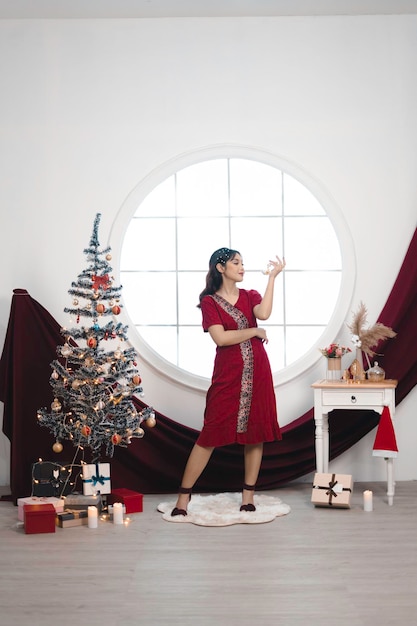 Retrato de una linda joven con un vestido rojo sonriendo a la cámara de pie en un salón de Navidad decorado en el interior