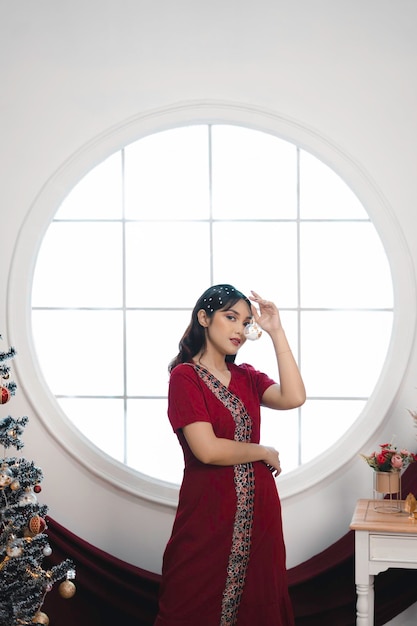 Retrato de una linda joven con un vestido rojo sonriendo a la cámara de pie en un salón de Navidad decorado en el interior