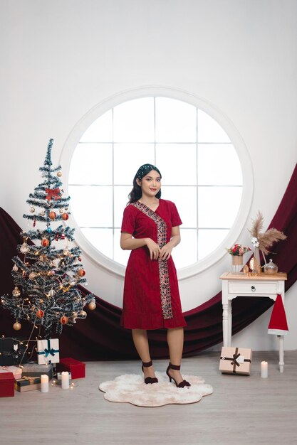 Retrato de una linda joven con un vestido rojo sonriendo a la cámara de pie en un salón de Navidad decorado en el interior