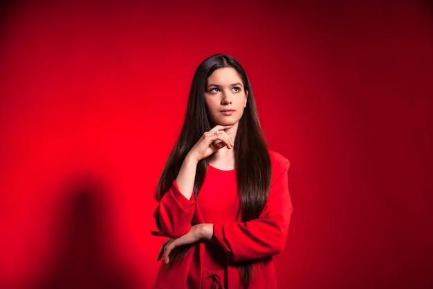 Retrato de una linda jovencita pensante con un elegante traje rojo sobre una pared de fondo roja mirando hacia otro lado