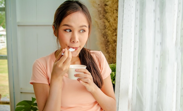 Foto retrato linda mujer asiática de pelo largo comiendo helado de leche dulce de una taza de pie junto a la ventana.