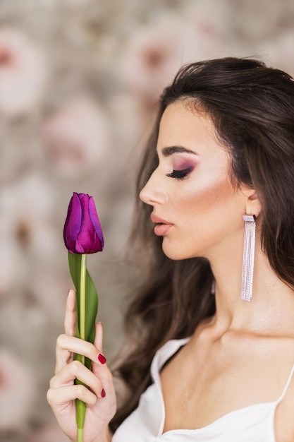 Un retrato de una linda mujer joven que sostiene un tulipán morado y representa un bonito maquillaje en su rostro.