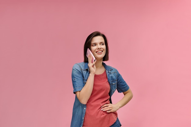 Retrato de linda mujer morena feliz vistiendo camiseta y jeans camisa hablando por teléfono móvil y sonriendo aislado sobre fondo de color rosa