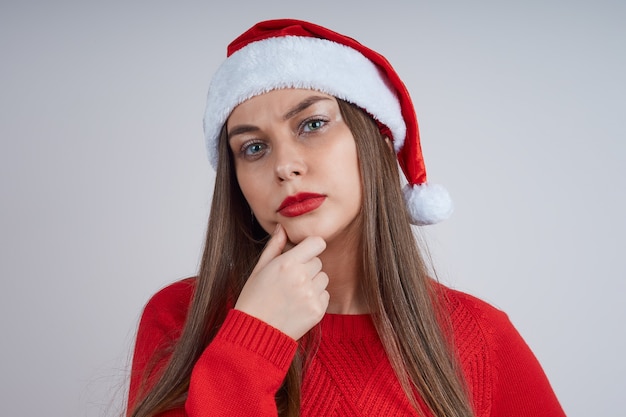 Retrato de linda mujer pensativa en suéter rojo y gorro de Papá Noel sobre fondo gris.
