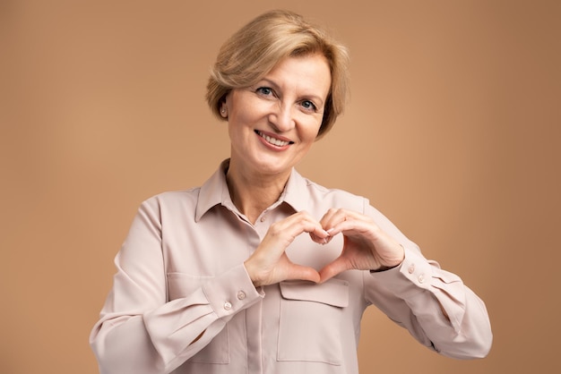 Retrato de linda mujer senior sonriente haciendo forma de corazón con las manos, expresando amor, amistad y cuidado, romance en la adolescencia. Tiro de estudio interior aislado sobre fondo beige