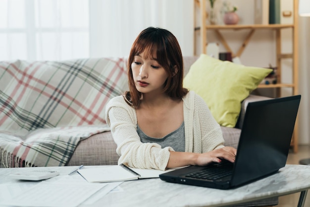 retrato linda mulher asiática está olhando para o documento financeiro e inserindo informações no laptop enquanto arquiva o imposto on-line na sala de estar em casa.