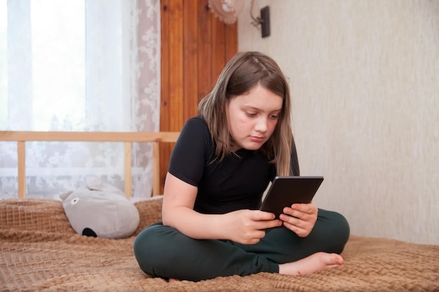 Retrato de una linda niña de 12 años con camiseta negra y pantalones de chándal en la cama con tableta