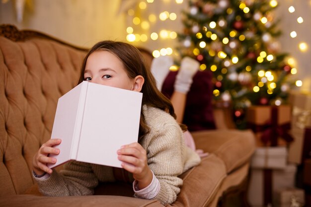 Retrato de linda niña acostada en el sofá y libro de lectura en la habitación decorada con árbol de Navidad