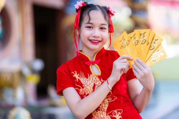 Retrato Linda niña asiática vistiendo rojo cheongsam chino tradicional, sosteniendo sobres amarillos con el texto chino