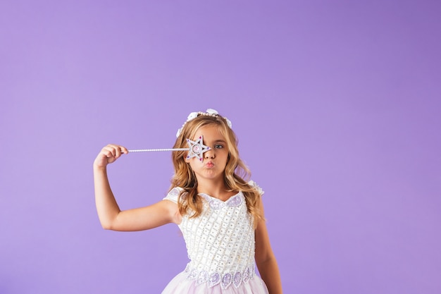 Retrato de una linda niña bonita sonriente vestida con un vestido de princesa aislado sobre pared violeta, sosteniendo la varita mágica