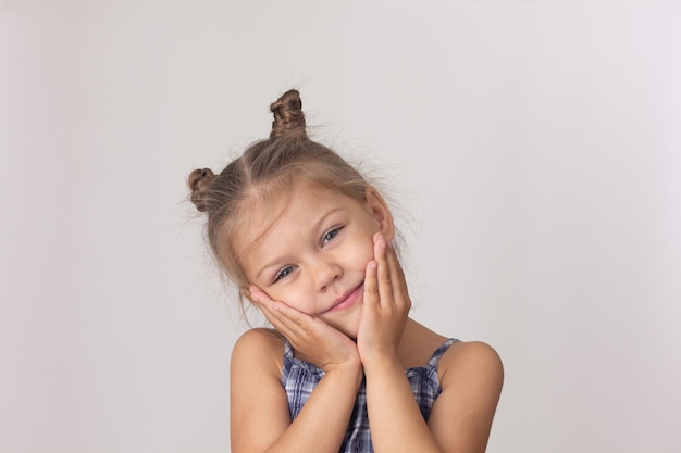 Retrato de una linda niña caucásica de cinco años sosteniendo las mejillas de la cabeza con las manos en el fondo blanco mirando a la cámara