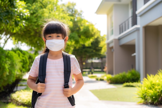Retrato de linda niña de escuela primaria con máscara médica frente a su casa durante la pandemia de COVID-19.