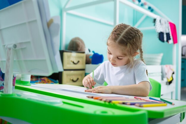 Retrato de una linda niña mirando a la cámara y sonriendo mientras dibuja o hace la tarea, sentada en una mesa en el interior de la casa, espacio de copia