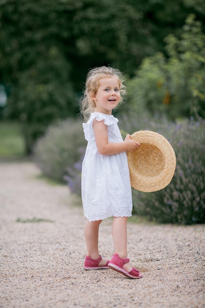 Retrato de una linda niña rizada de ojos azules de 3 años con un sombrero de mimbre y un vestido blanco cerca de un niño feliz de lavanda floreciente en la naturaleza
