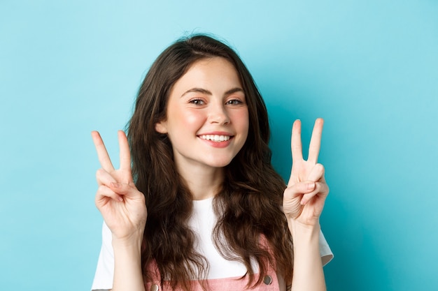 Retrato de linda niña sonriente mostrando signos de paz y mirando feliz a la cámara, de pie en traje de primavera sobre fondo azul.