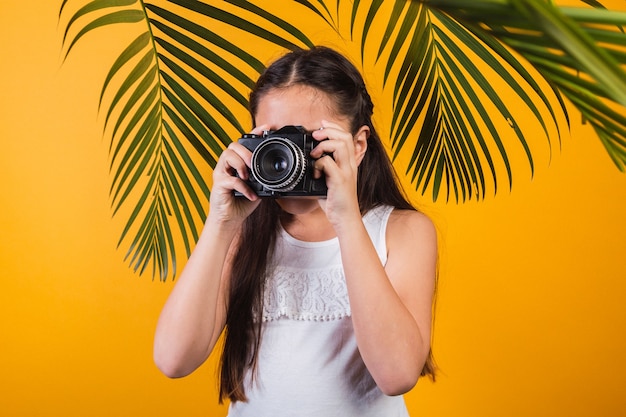 Retrato de una linda niña tomando fotografías con una cámara sobre fondo amarillo.