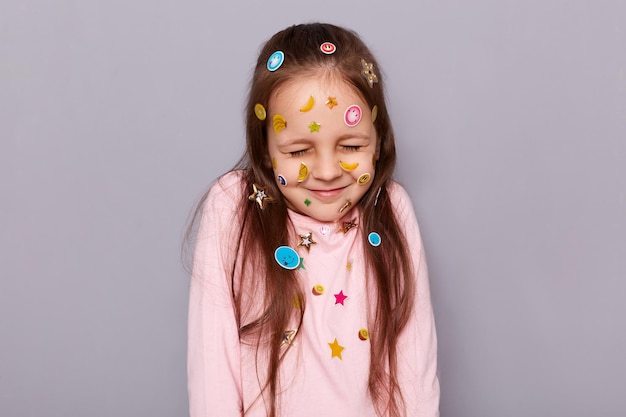 Retrato de una linda niñita de cabello castaño soñando cubierta con pegatinas posando aislada sobre fondo gris parece tímida con los ojos cerrados y sonriendo