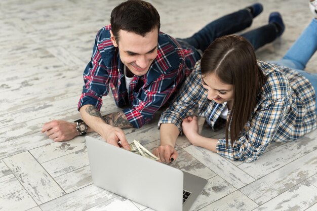 Foto retrato de una linda pareja joven tendida en el suelo en casa y administrando el presupuesto usando una computadora portátil