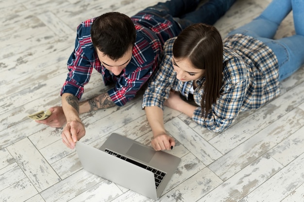 Foto retrato de una linda pareja joven tendida en el suelo en casa y administrar el presupuesto usando la computadora portátil