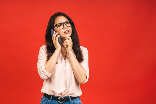 Retrato de una linda y seria mujer de negocios joven y alegre que usa una conversación informal por teléfono móvil aislada sobre un fondo rojo