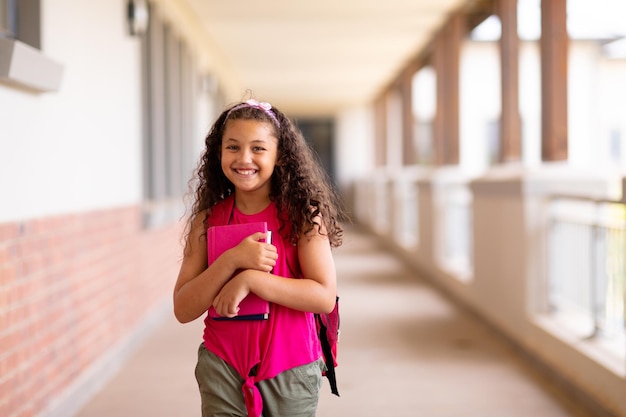 Retrato de una linda y sonriente chica birracial sosteniendo libros mientras estaba de pie en el pasillo de la escuela