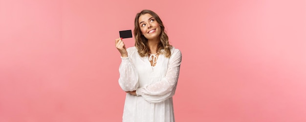 Retrato de una linda y tierna chica rubia femenina soñando despierta con cómo desperdiciará su dinero en compras en línea, busque imágenes de ensueño mientras espera la entrega, sostenga el fondo rosa de la tarjeta de crédito