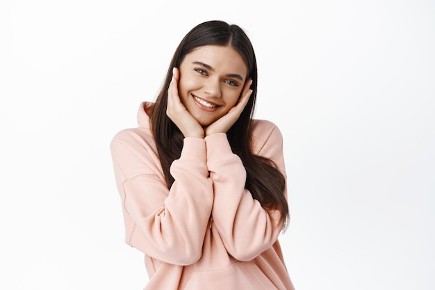 Foto retrato de linda y tierna niña morena sonrojada por una noticia feliz o sorpresa, tomados de la mano en las mejillas y sonriendo, de pie contra la pared blanca