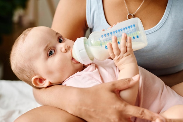 Foto retrato de un lindo bebé mirando la cámara mientras come leche del biberón y está acostado en los brazos de su madre