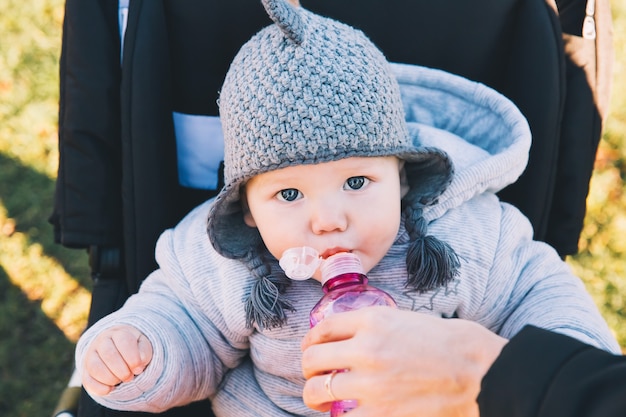 Retrato de un lindo bebé en un paseo al aire libre. El niño se sienta en un cochecito y bebe agua de la botella.